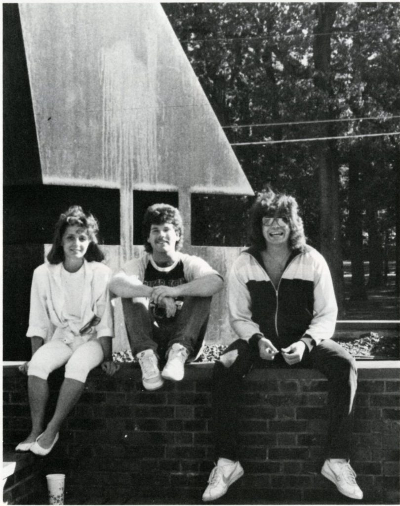 Three students, perhaps in the 1970s, sit outside the Student Center in front of a sculpture. 