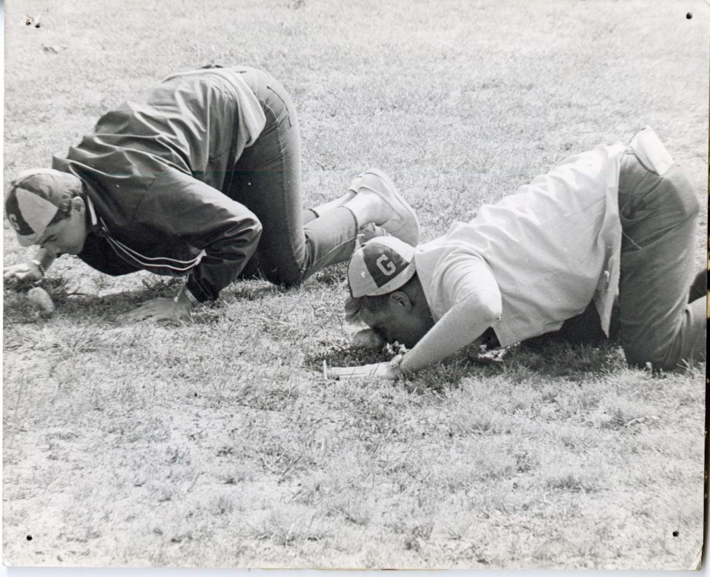 Two male students wearing Glassboro hats push potatoes with their noses, crouched on the ground. 