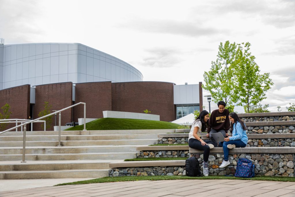 Three students in 2023 sit on the amplitheatre steps that replaced the bench, with Wilson Hall behind them. 