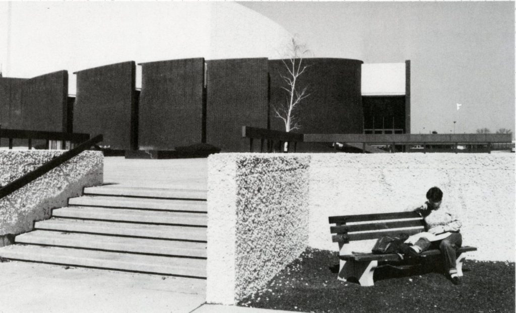 A student sits on a bench with Wilson Hall behind them. Perhaps the 1970s. 