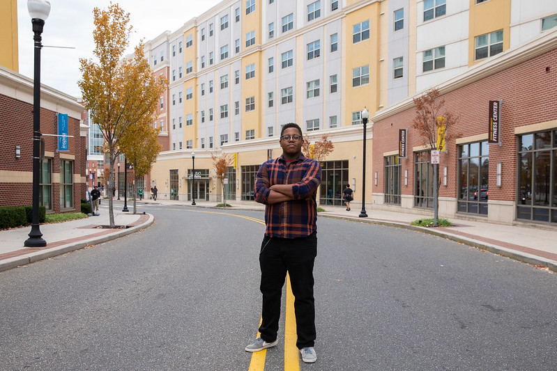Sedrick stands in a serious manner, with arms crossed, on the yellow lines in the middle of a quiet street in Glassboro. 