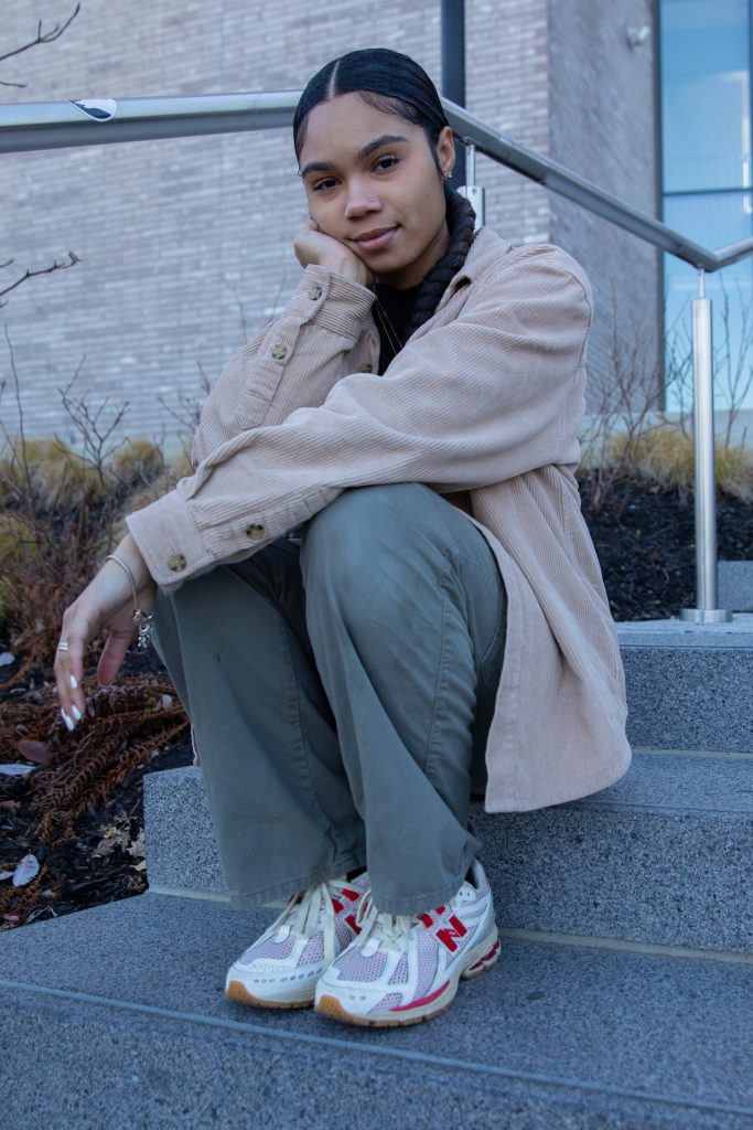 Student Dabany Garris sitting on the steps in front of Discovery Hall. She can be seen in a tan jacket and green cargo pants.