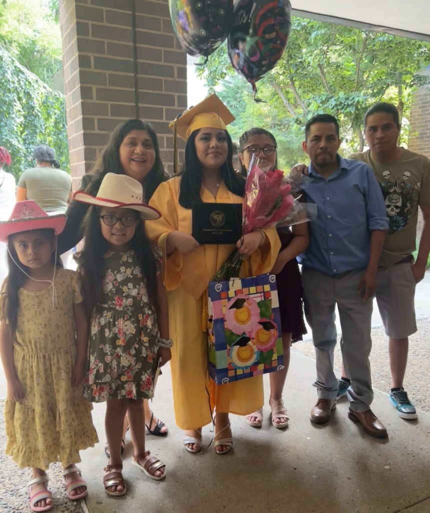 Rosa stands surrounded by family after her high school graduation, wearing a golden yellow graduation gown. 